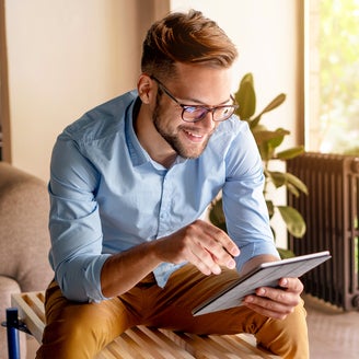 The picture shows a young man with glasses, leaning slightly forwards and sitting on a modern wooden bench. The bench is in a bright and cosy room. The man is wearing a light blue shirt and mustard-coloured trousers. He is holding a tablet in his hands and looking at ista's heat metering system. Behind the man, to the right of the picture, is a large floor-to-ceiling window that allows plenty of natural light into the room. A green garden with lush vegetation can be seen through the window. Next to the window is a large plant in a pot on the floor. To the right of the plant is a black radiator, which contrasts with the light and natural colours of the room. To the left of the man, part of a beige sofa is visible. Other parts of the room can be seen in the background on the left of the picture, possibly including a door or another window, which also bring light and space into the room.